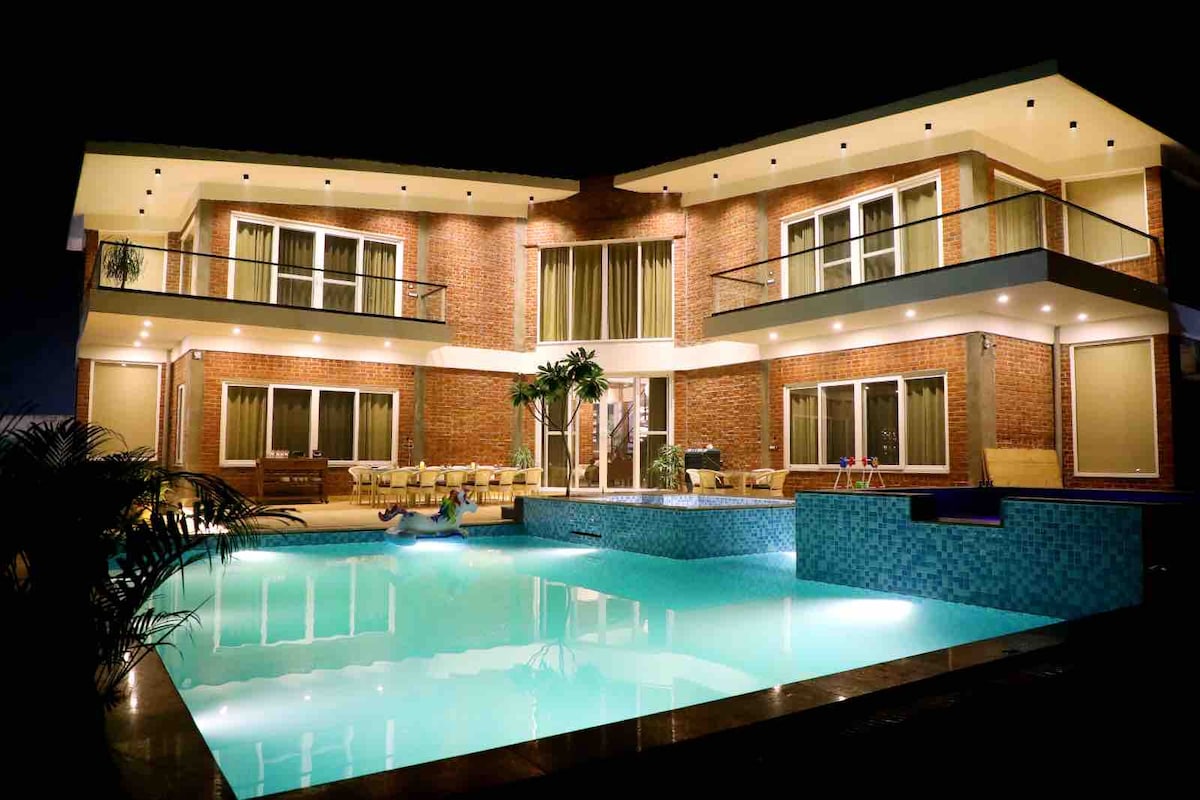 The property is two stories high, with exposed brick walls creating a warm ambiance. The private infinity pool reflects the building's lighting, surrounded by lounge seating. Floor-to-ceiling windows enhance the sense of spaciousness, offering views of the pool area from multiple angles.