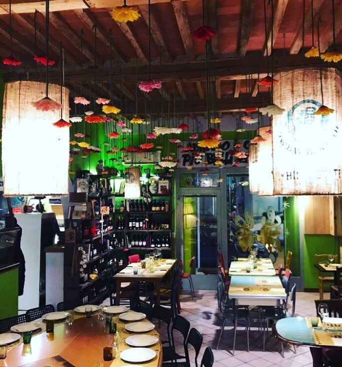 The dining area features an array of hanging decorations above wooden tables set with plates and glasses. Shelves lined with bottles create an inviting backdrop. Warm lighting enhances the vibrant atmosphere, while colorful floral accents add a lively touch to the space.