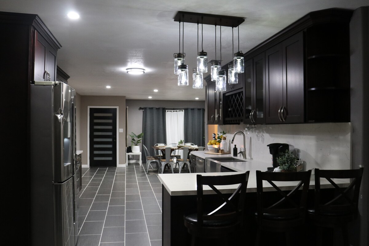 The modern kitchen features dark cabinetry and a spacious island with seating. A set of glass pendant lights enhances the workspace, while stainless steel appliances and a coffee maker are included. A dining area is visible in the background, accentuated by natural light from the nearby window.