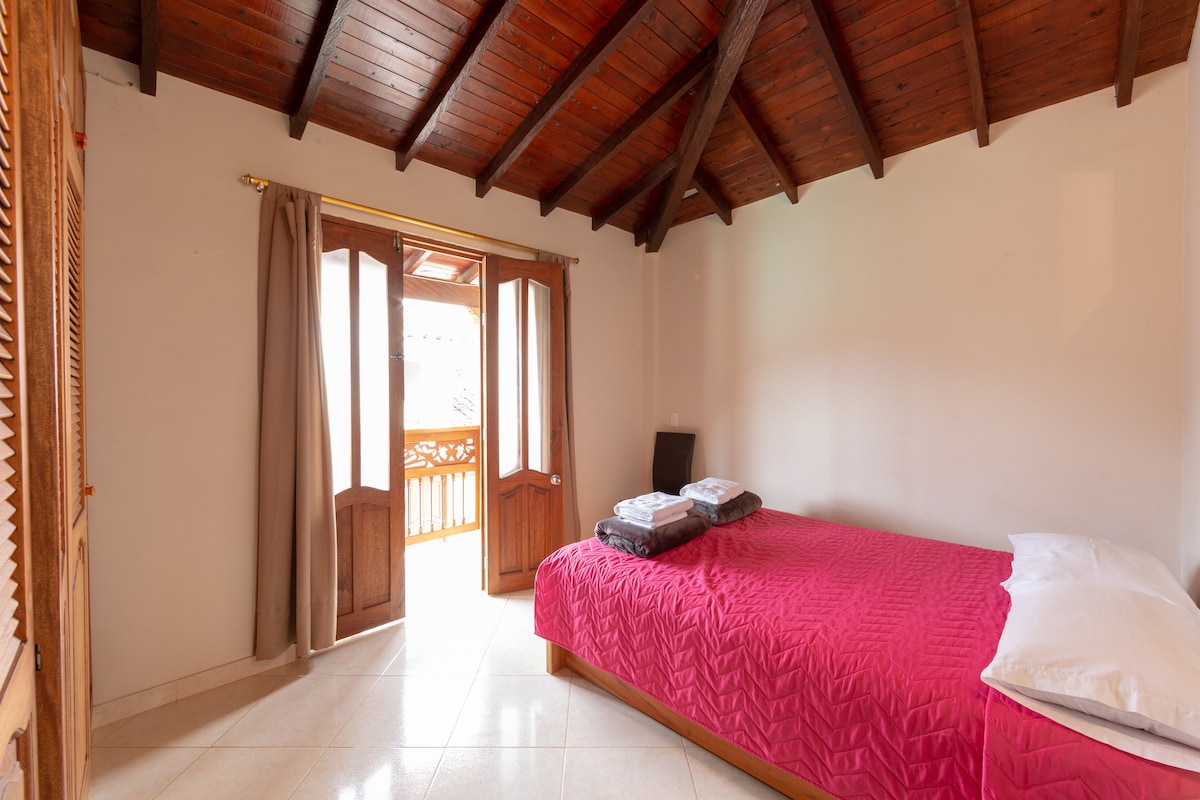 A bright room features a spacious bed with a quilted red coverlet and white pillows. Two wooden doors lead to a balcony, allowing natural light to fill the space. Fresh towels are neatly arranged at the foot of the bed against the light-colored walls.
