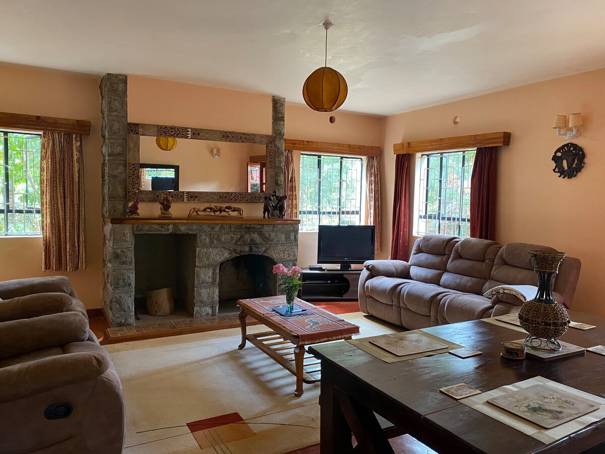 A spacious living area features a stone fireplace as a focal point, with comfortable seating arranged around a central coffee table. Large windows allow natural light to brighten the room, complemented by soft curtains. A television is positioned nearby, providing an inviting atmosphere for relaxation.