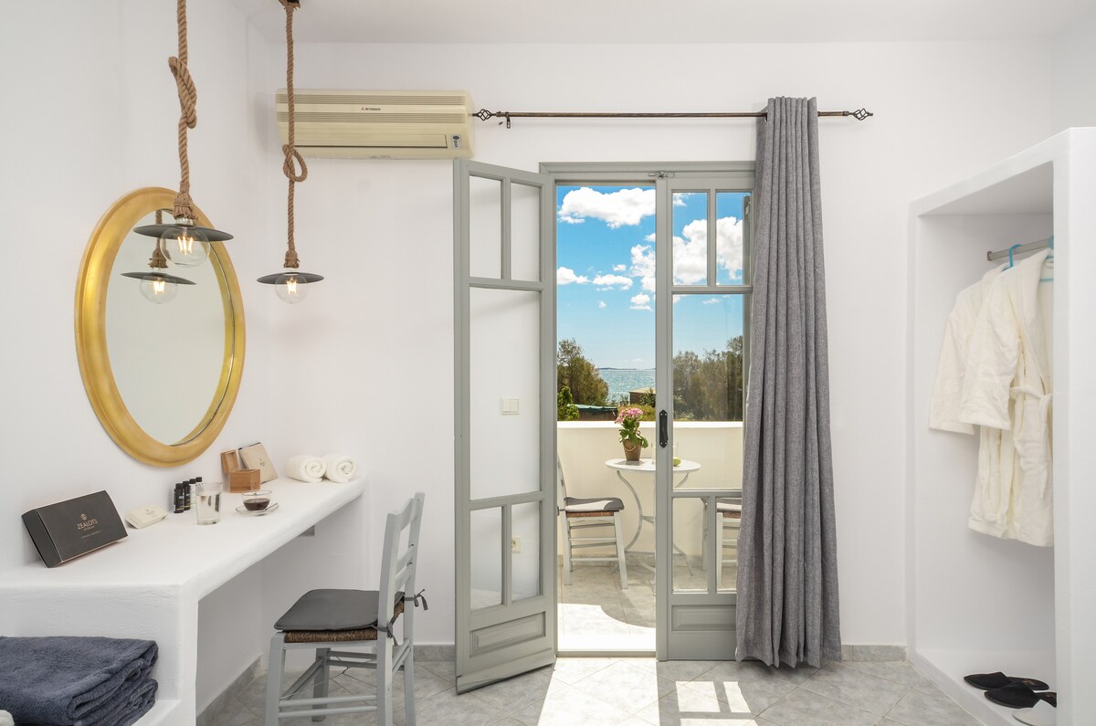 A bright room features double doors leading to a small balcony with scenic views. A round mirror reflects natural light. A desk with a chair is positioned beside a shelf holding neatly arranged towels and bathrobes.