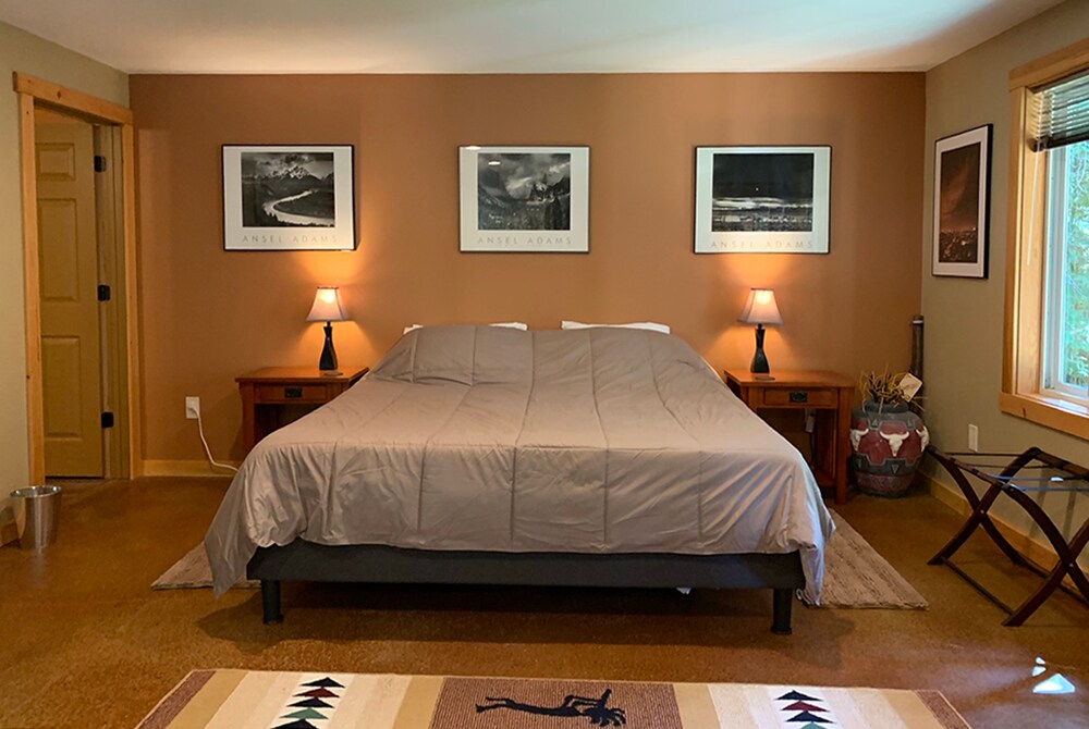 A spacious open layout bedroom features a king-size bed with a neutral-colored blanket. Two bedside tables with lamps are positioned next to the bed, while framed black-and-white photographs adorn the walls. Natural light filters through a window, enhancing the warm tones of the room.
