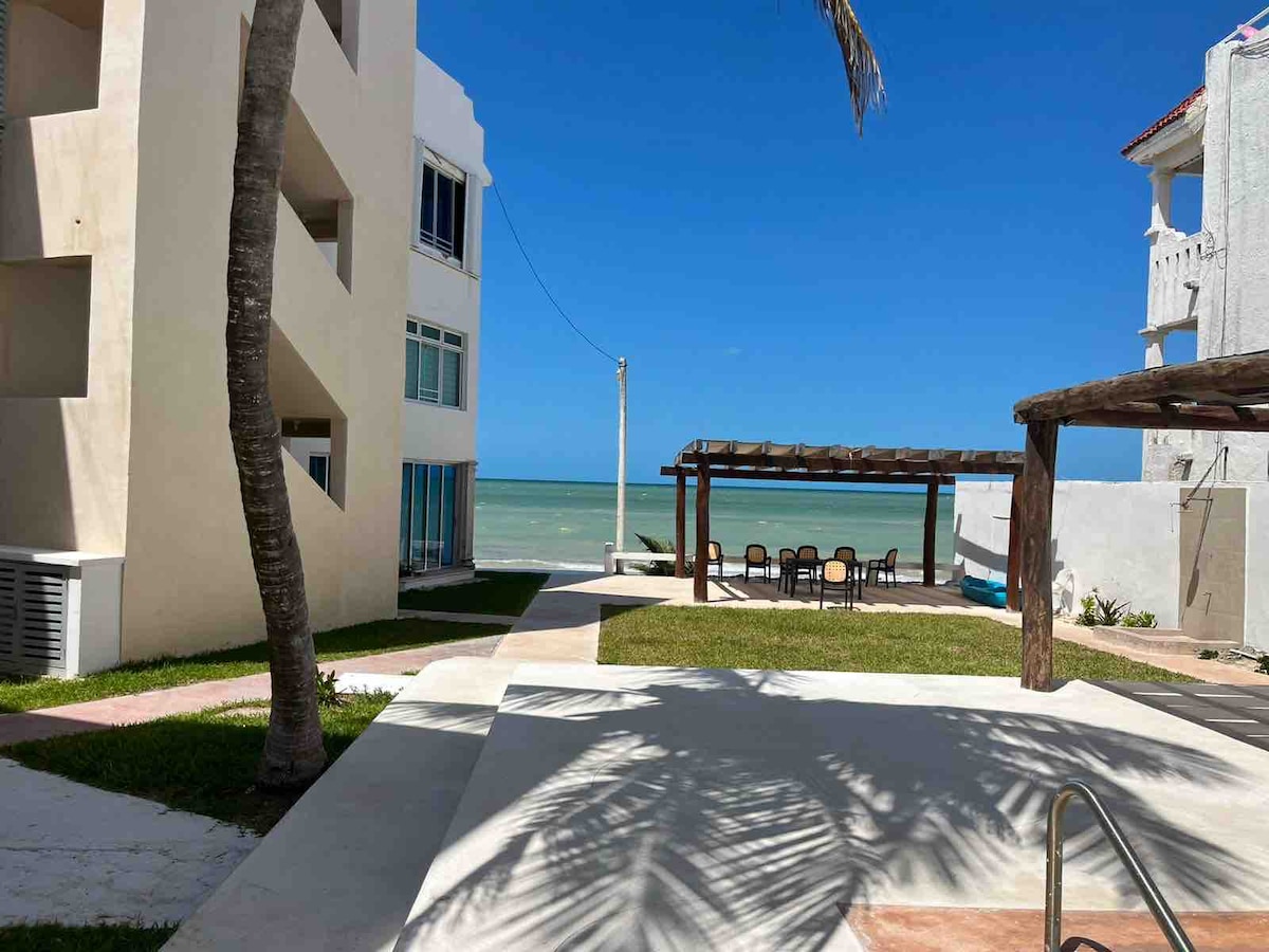 A well-maintained outdoor area features a pathway lined with grass and palm trees, leading to a comfortable seating arrangement under a wooden pergola. The clear blue sky and serene sea create a tranquil backdrop, with several buildings visible in the surrounding landscape.