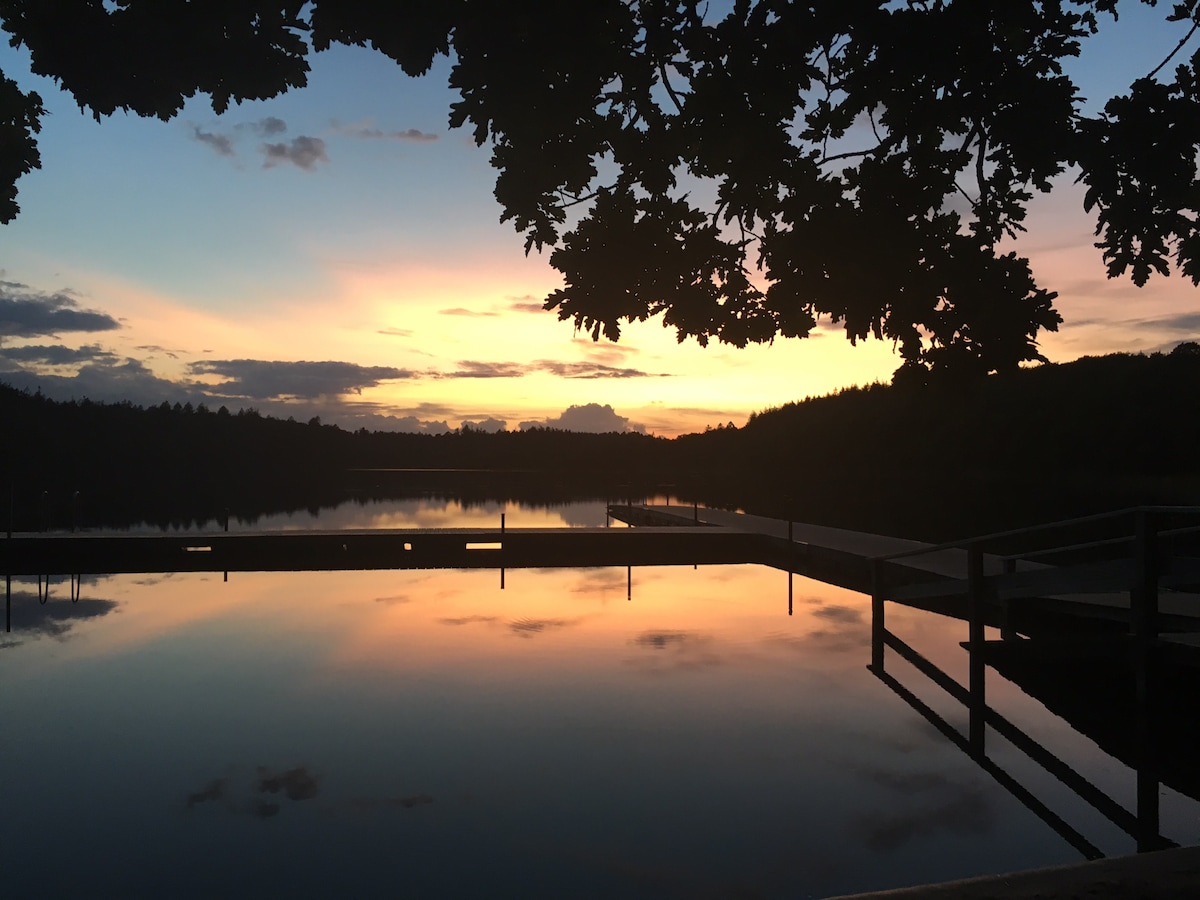 A serene lakeside scene is captured during sunset, with vibrant colors reflecting off the calm water. Silhouettes of trees frame the image, and wooden docks extend along the shoreline, providing a tranquil setting for relaxation.