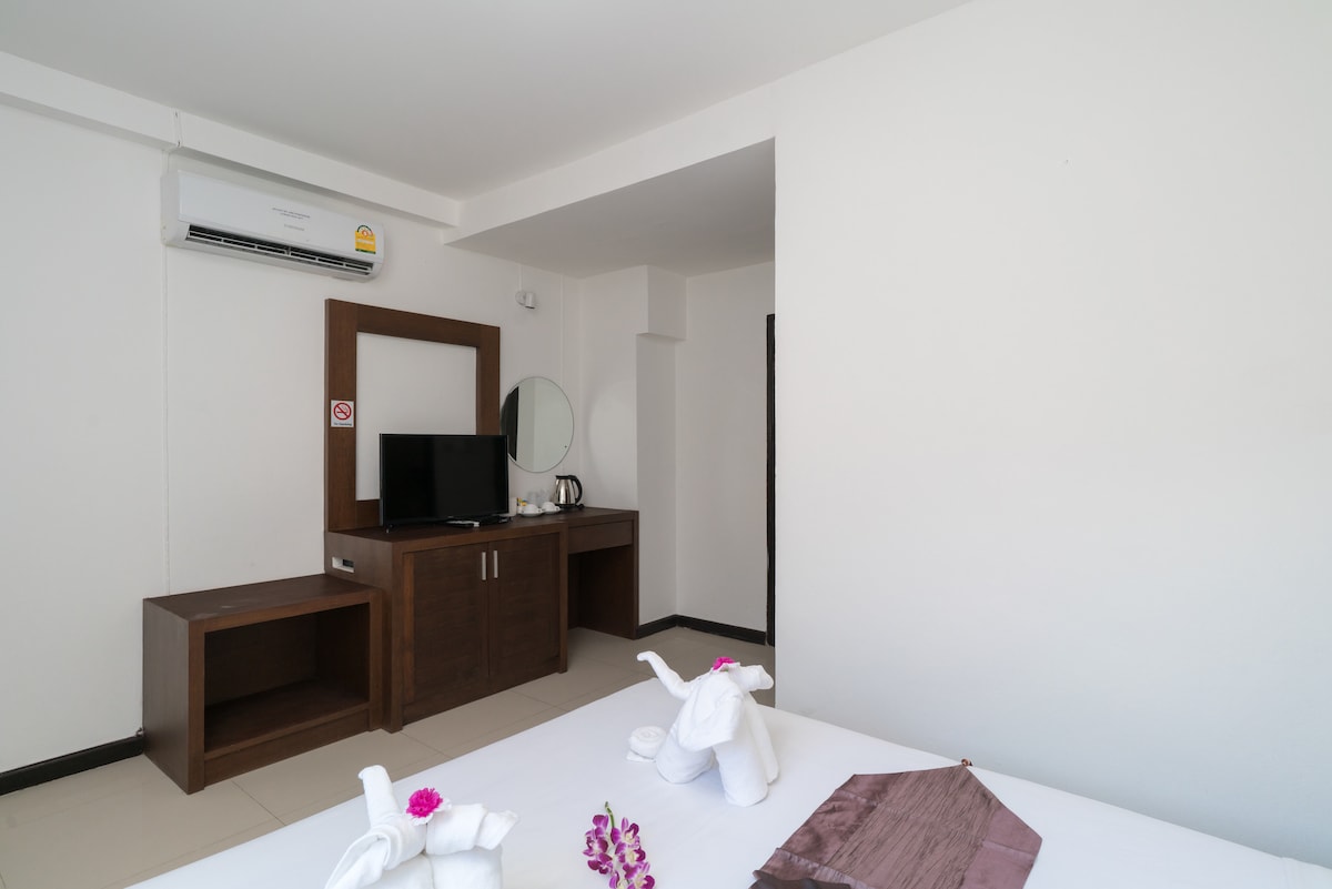The room features a queen-size bed adorned with fresh towels shaped like elephants. A modern flat-screen TV is positioned on a wooden console, alongside an electric kettle. A round mirror reflects light, complementing the air-conditioning unit installed above.