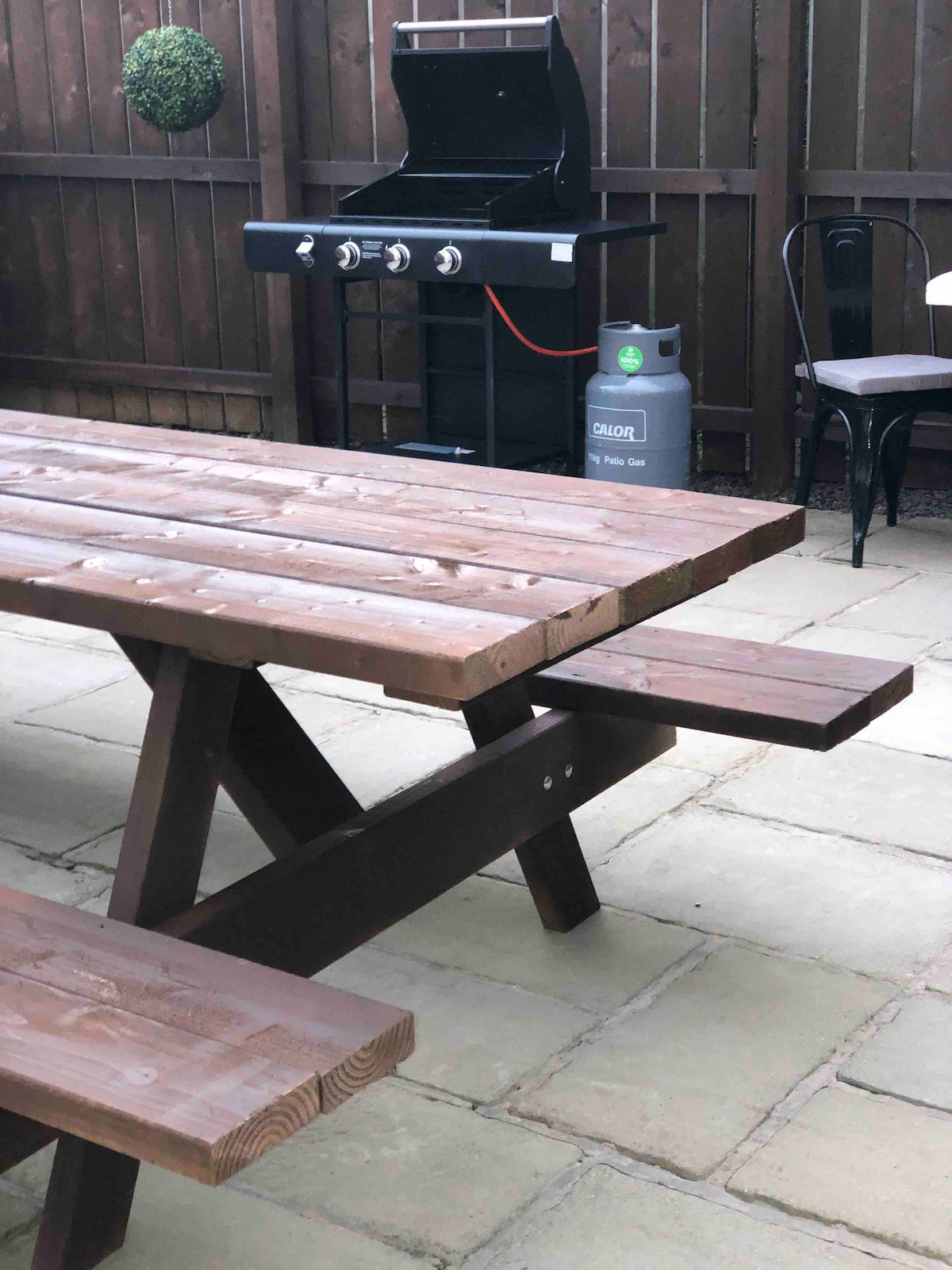 A spacious paved garden features a wooden picnic table with benches and a black gas grill positioned against a wooden fence. A propane tank is visible nearby, complementing the outdoor cooking space.