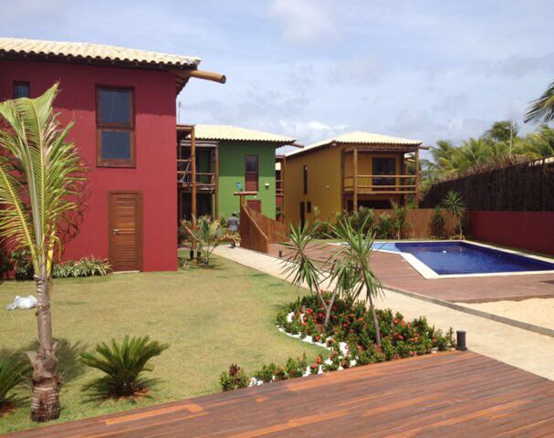 A landscaped outdoor area features a well-maintained garden with palm trees and colorful flowers. Two houses in vibrant colors are visible, along with a clear blue swimming pool. A wooden deck offers a space for relaxation, surrounded by lush greenery.