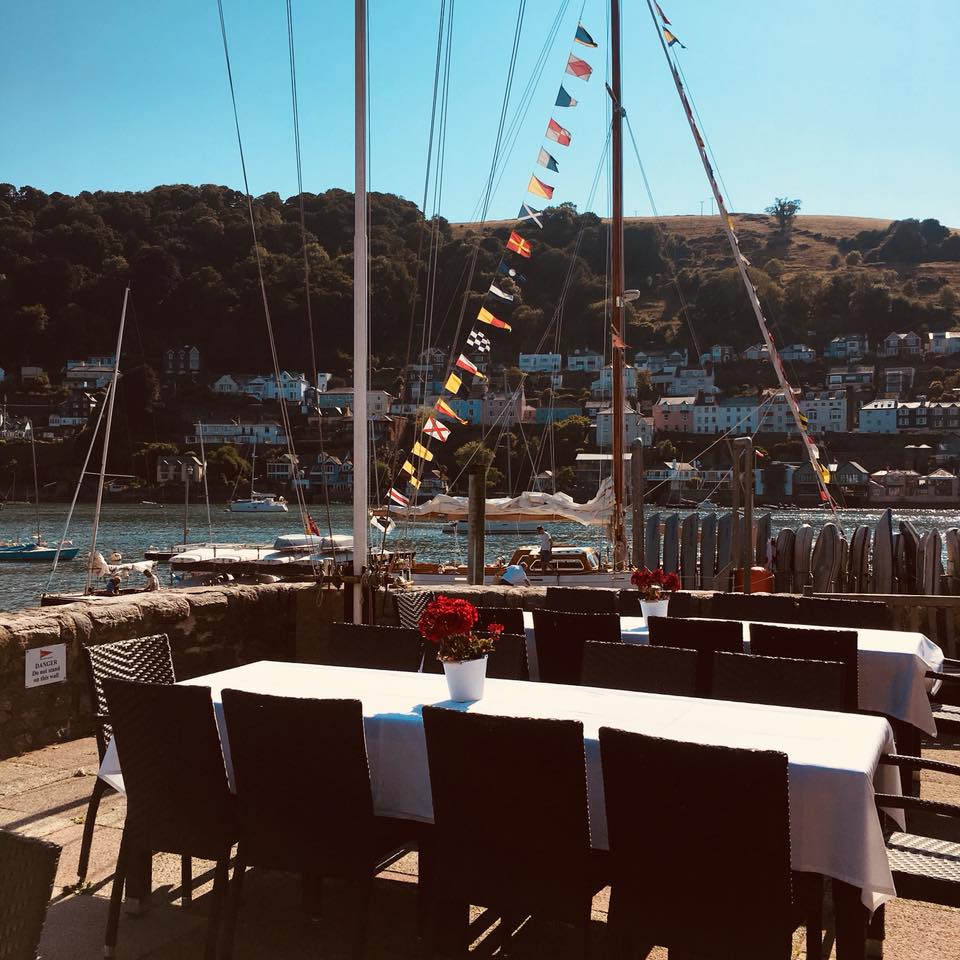 An outdoor terrace features a row of black wicker tables set with white tablecloths, complemented by vibrant red flowerpots. The view includes a lively waterfront scene with boats sailing and colorful flags fluttering in the breeze against a backdrop of green hills and houses.