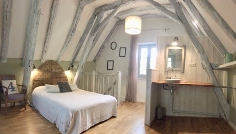 The bedroom features a spacious bed with a light bedding set, framed by exposed wooden beams that create an airy atmosphere. A small kitchenette is present in one corner, complemented by natural light from the nearby window. Soft-toned decor enhances the room's calming nature.