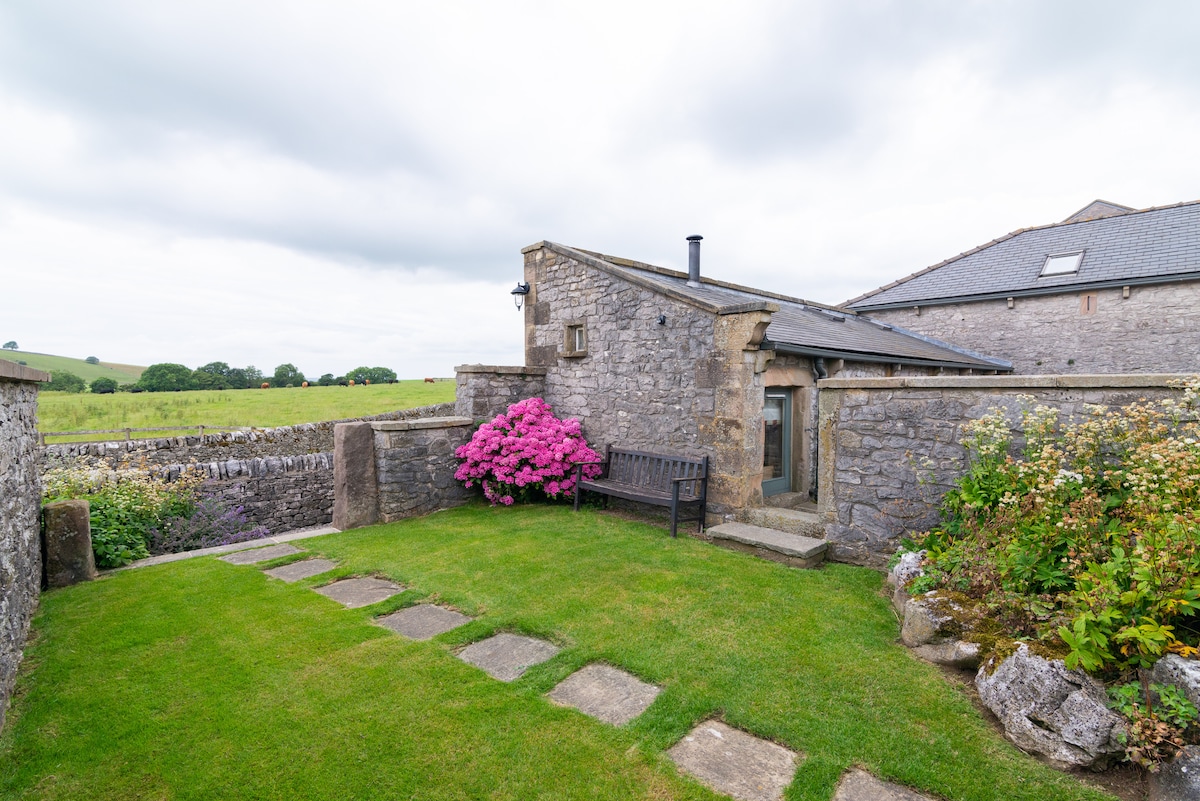 A stone cottage entrance is framed by vibrant pink flowers and greenery. A pathway of stone slabs leads through a well-maintained lawn area, with a bench positioned against a stone wall. Open countryside extends in the background under an overcast sky.