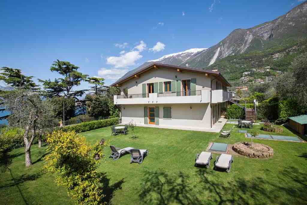The building showcases a two-story structure surrounded by a lush green garden. Comfortable lounge chairs and sunbeds are placed on the lawn, with a seating area highlighted by a round table. A picturesque mountain backdrop complements the serene environment.