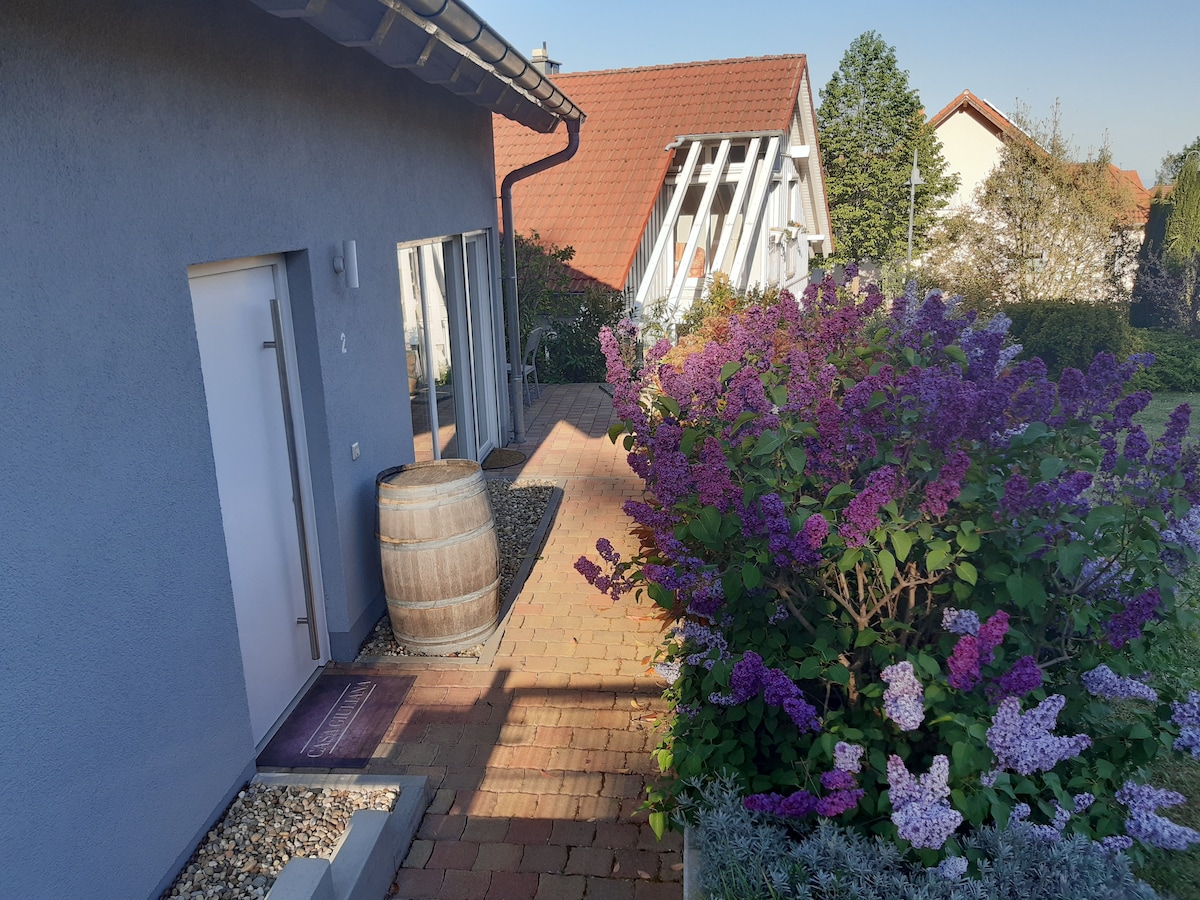A peaceful pathway lined with blooming lilacs leads to the entrance of the house. A decorative wooden barrel is visible, complementing the exterior's subtle hues and the natural surroundings. Large windows allow ample light, contributing to a welcoming atmosphere.