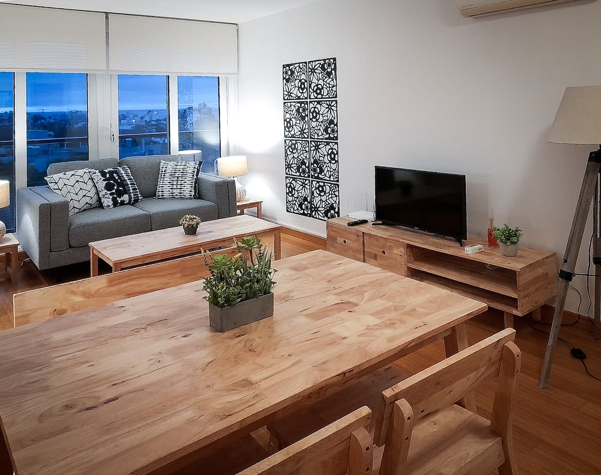 The living area features a spacious sofa, a wooden coffee table, and a dining table surrounded by chairs. Natural light fills the room through large windows, enhancing the simple decor that includes a patterned wall art piece and modern lighting.