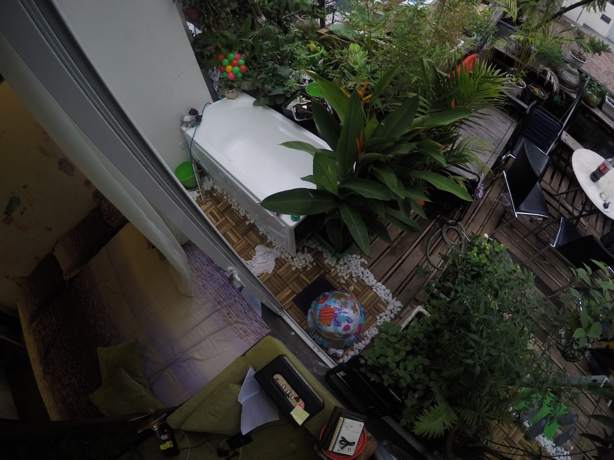 An inviting outdoor space is surrounded by lush greenery, featuring a large bathtub among vibrant plants. A wooden deck is visible with a mix of seating options. Colors from decorative items and plants create a serene atmosphere, ideal for relaxation in a private setting.