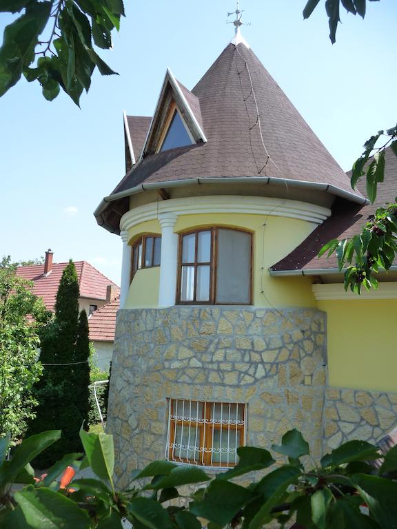 A charming three-story tower apartment is viewed from a distance, showcasing a stone base and a sloped, shingled roof. Bright yellow walls are complemented by large windows, framed by white columns. Surrounding greenery partially obscures the view, creating a serene atmosphere.
