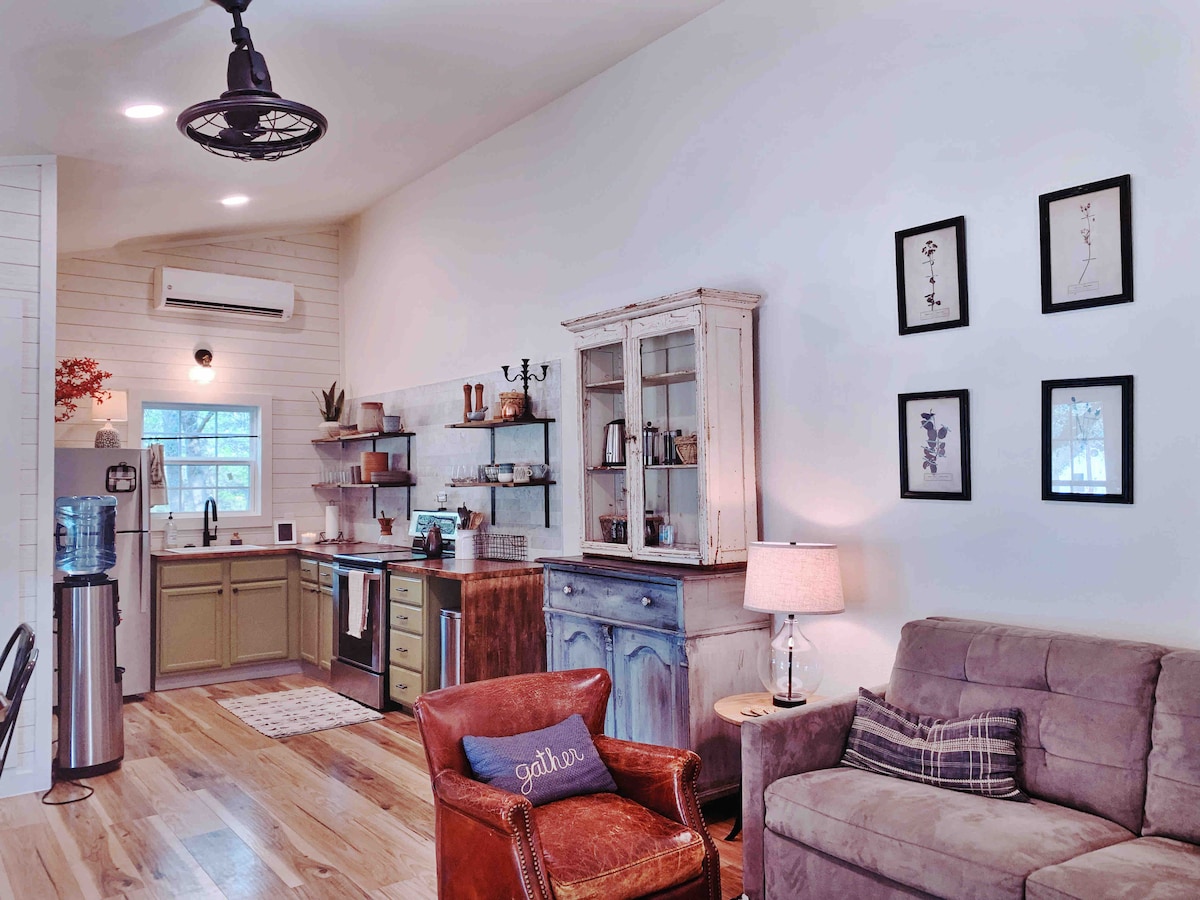 A welcoming living area features a comfortable sofa and an armchair, accented by soft lighting from a side table lamp. The kitchen area, equipped with modern appliances, showcases rustic shelving and a vintage hutch, adding character to the space. Natural light flows through the window.