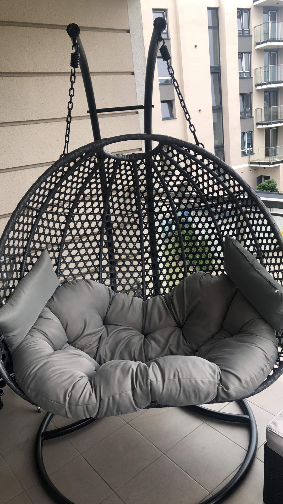 A cozy hanging chair is featured on the balcony, characterized by a woven design and plush gray cushions. The chair is suspended from a sturdy frame, creating a comfortable lounging area with views of the surrounding buildings and greenery.