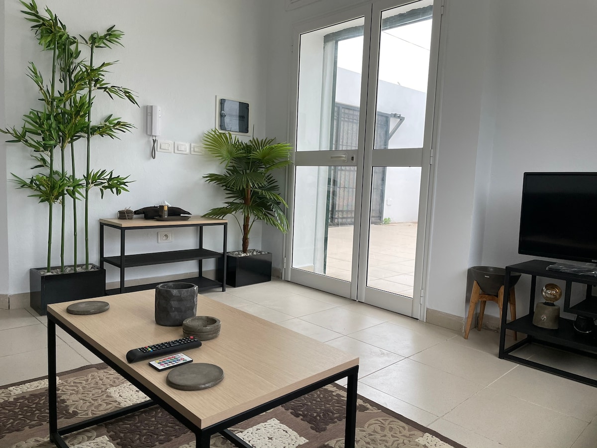 A living area is presented with a light-colored coffee table, complemented by decorative plates and a remote control. Tall green plants add a natural touch, while large glass doors lead to an outdoor space. A television is positioned on a sleek stand.