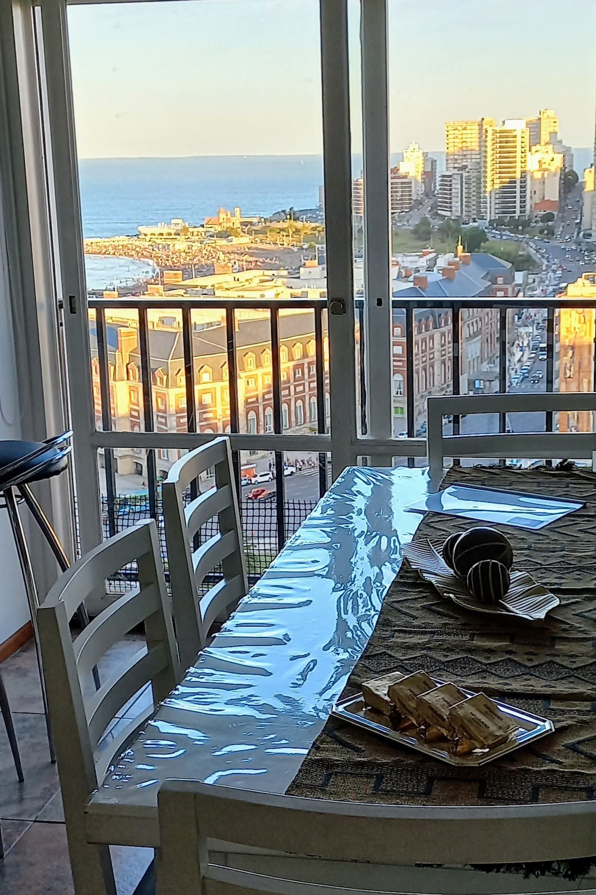 A dining area is presented with a clear table set for four, featuring decorative elements and a table runner. Expansive windows frame a view of the ocean and the city skyline, with warm sunlight casting a glow over the scene.