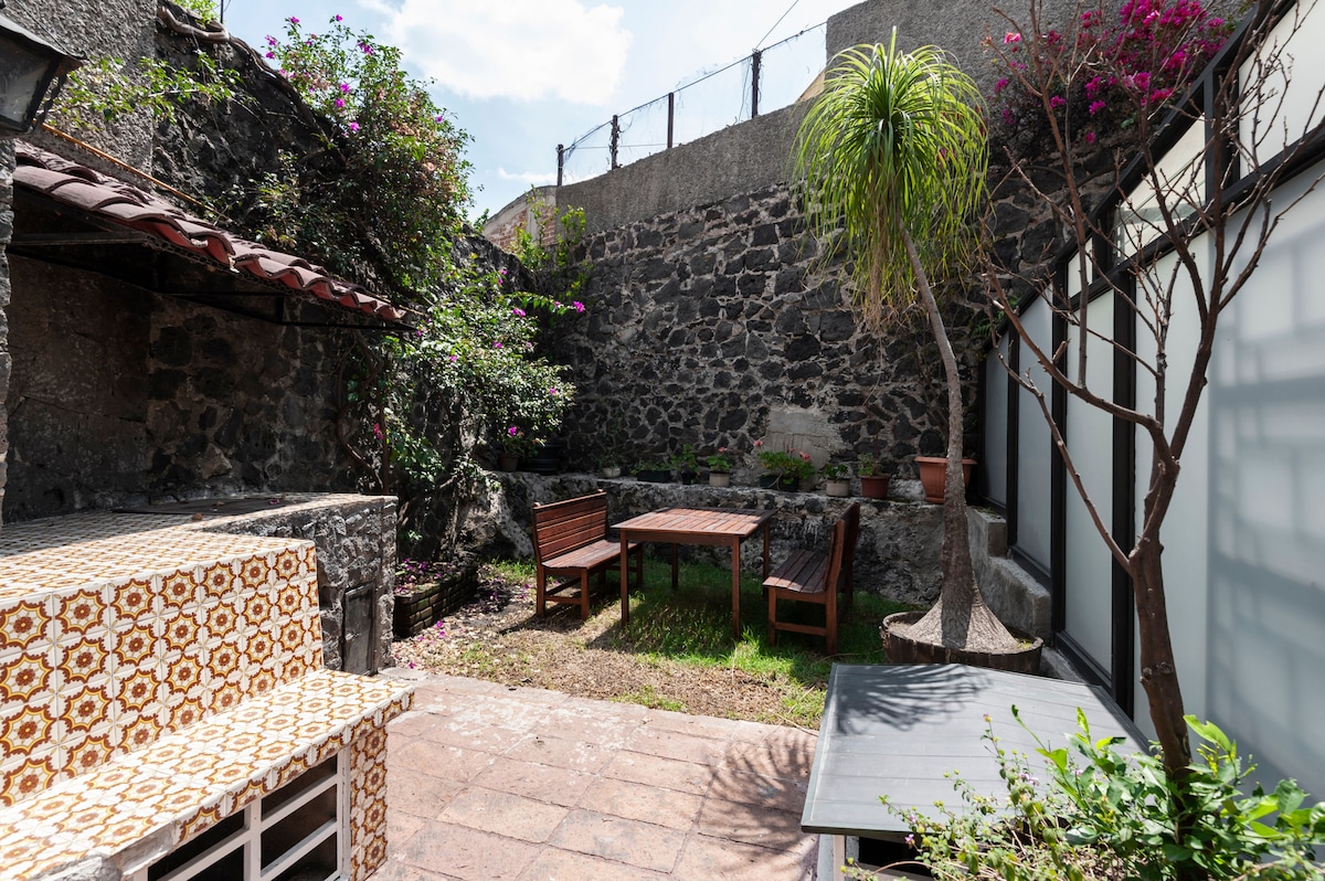 A private garden space is presented, featuring a stone wall adorned with vibrant flowering plants. A wooden dining table accompanied by benches occupies the center, with a patio area showcasing a patterned seating structure. Sunlight filters down, illuminating the serene outdoor setting.