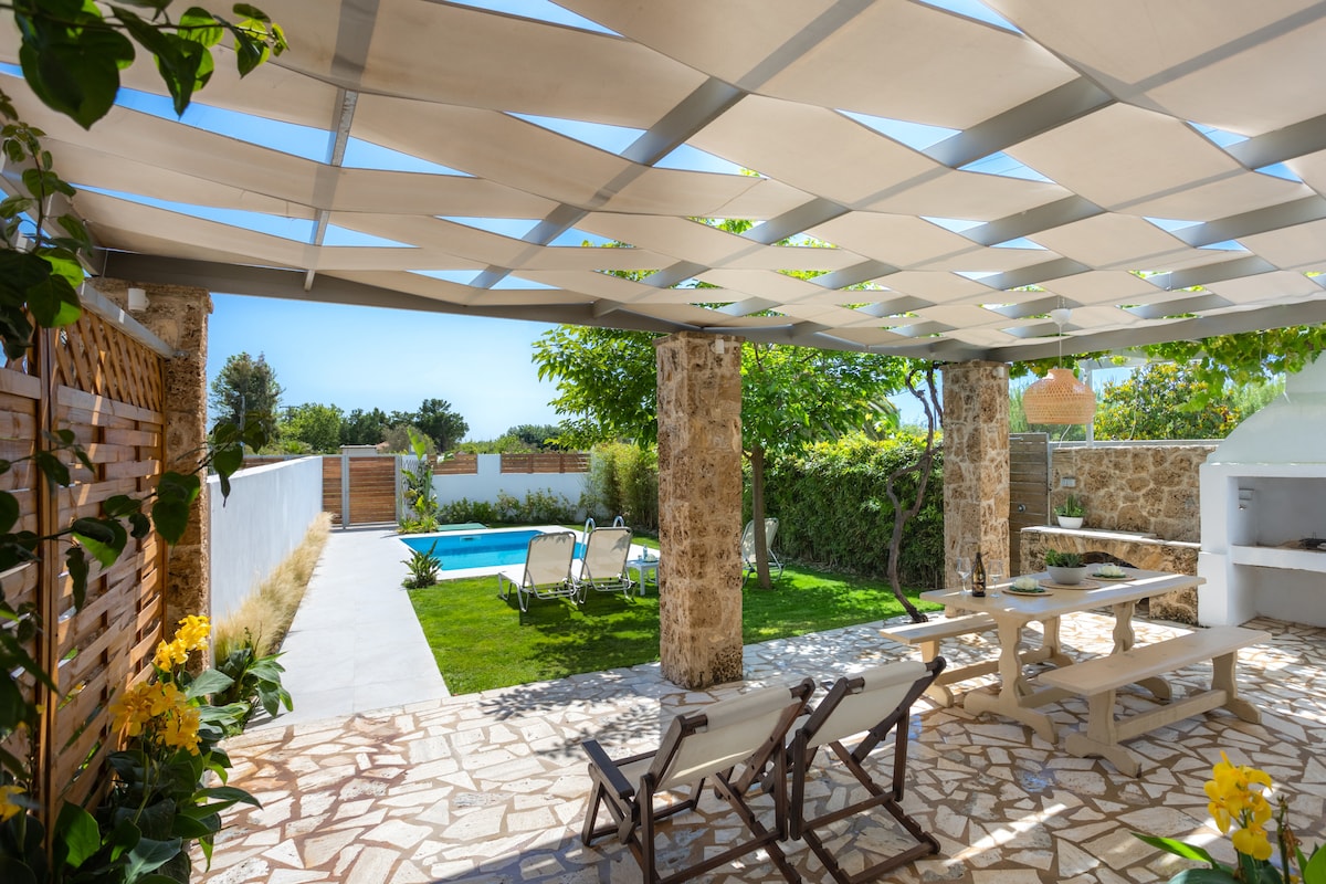 An outdoor patio area features stone flooring and shaded seating arrangements under a trellis. A dining table is situated nearby, accompanied by lounge chairs. In the background, a swimming pool is visible, surrounded by greenery, creating a serene atmosphere.