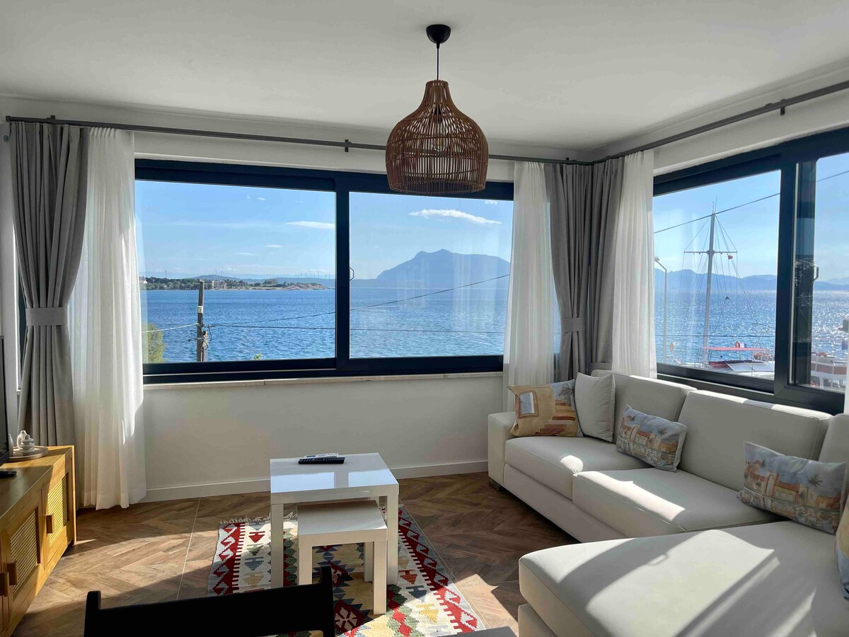 A bright living area is highlighted by large windows offering views of the sea and distant mountains. A light-colored sofa is paired with a small coffee table, while natural light enhances the space through elegant sheer curtains. Decorative pillows add subtle color to the seating.