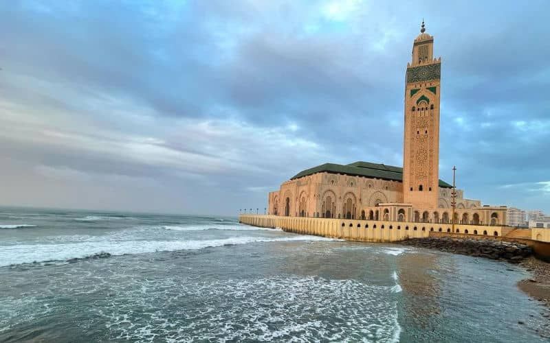 The image shows the exterior of the Hassan II Mosque, characterized by intricate architectural details and a tall minaret. Waves gently lap against the stone structure, with a backdrop of cloudy skies creating a serene coastal atmosphere.