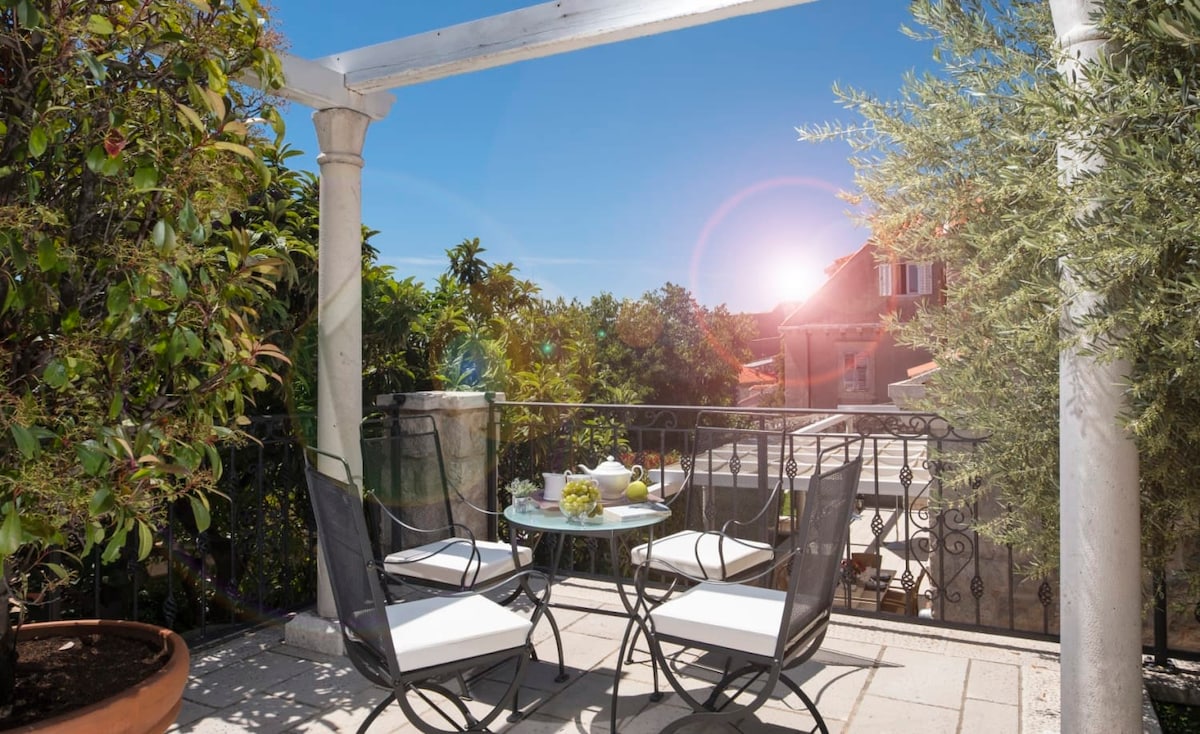 An outdoor terrace is framed by lush greenery and features a glass table with four metal chairs. The morning sun casts a warm glow, enhancing the peaceful ambiance. A view of surrounding rooftops and trees can be seen in the background.