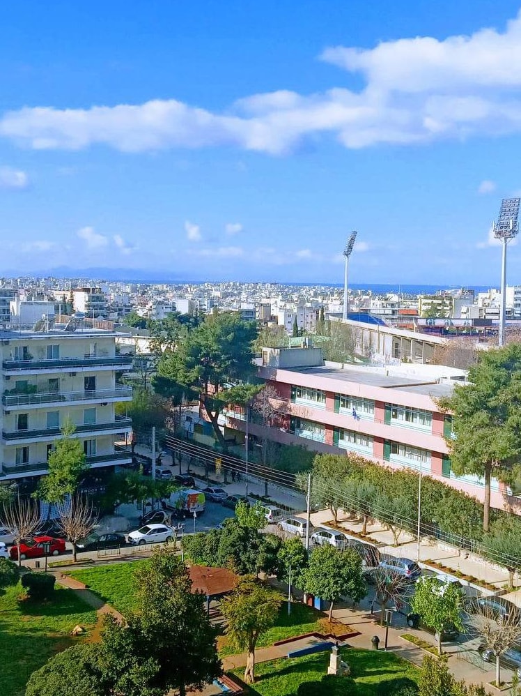 A panoramic view of the cityscape is captured, featuring buildings and greenery below. The horizon reveals the coastline, with a clear blue sky above and distant hills. A park is visible in the foreground, framed by trees and pathways.
