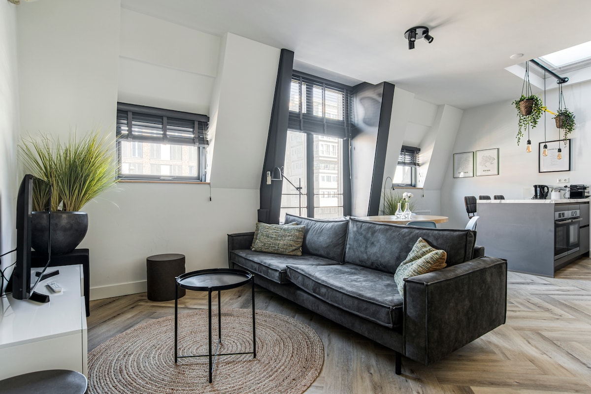 A modern living area is highlighted by a dark grey sofa and a small round table with a woven rug underneath. Natural light enters through large windows adorned with black blinds. The open kitchen is visible in the background, featuring contemporary appliances and décor.