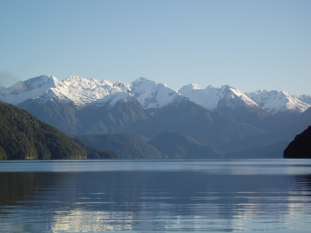 The tranquil surface of Lake Manapouri reflects the surrounding mountains, which are capped with snow. Lush green hills frame the lake, contrasting with the blue sky that stretches overhead. The peaceful scene invites relaxation and appreciation of the natural beauty.