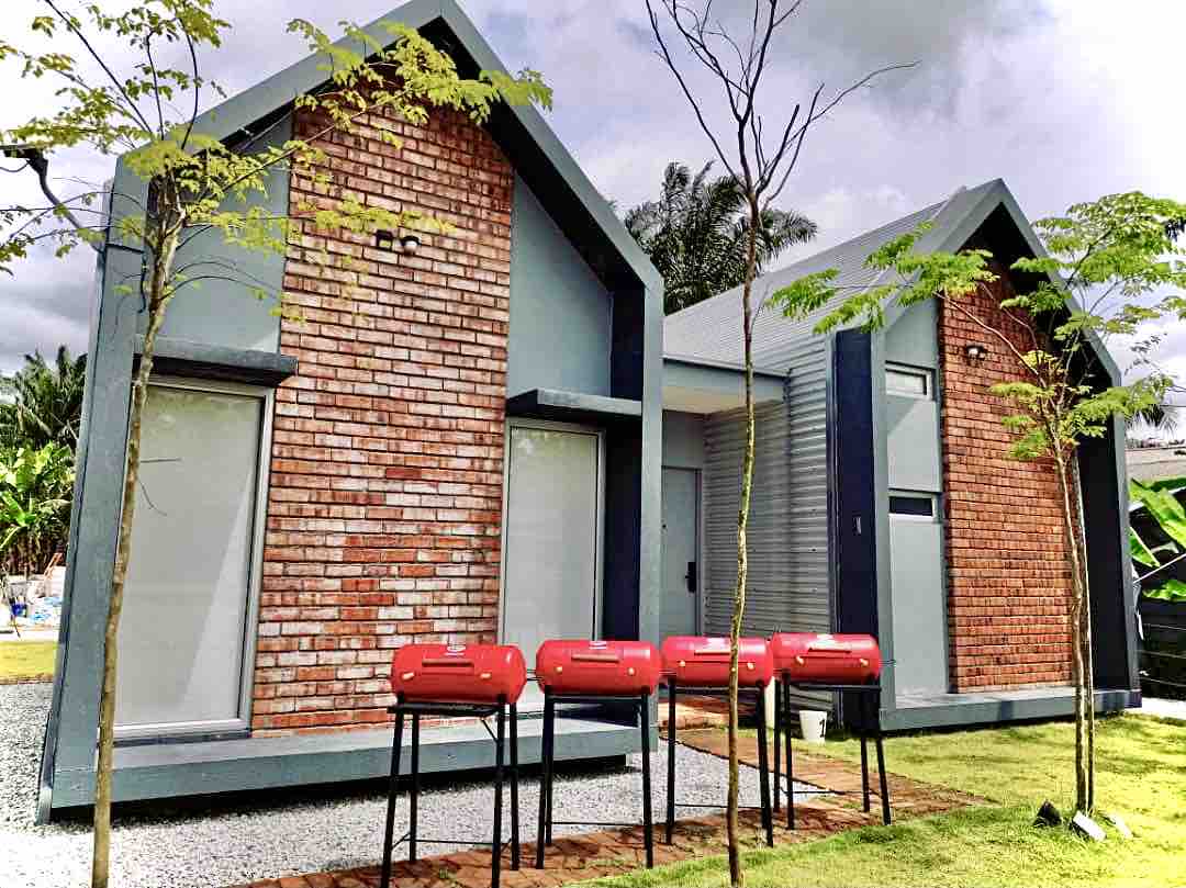 Two modern studio units are shown, featuring a blend of brick and grey exteriors. In the foreground, four red BBQ grills are placed on black frames, complemented by patches of green grass and small trees. Large windows provide a clear view of the inviting interiors.