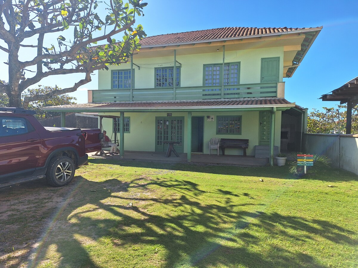 The two-story house features a vibrant green exterior with blue window shutters. A spacious lawn surrounds the property, complemented by a large tree casting shadows on the grass. Various outdoor seating areas are visible, offering a welcoming space for gatherings.