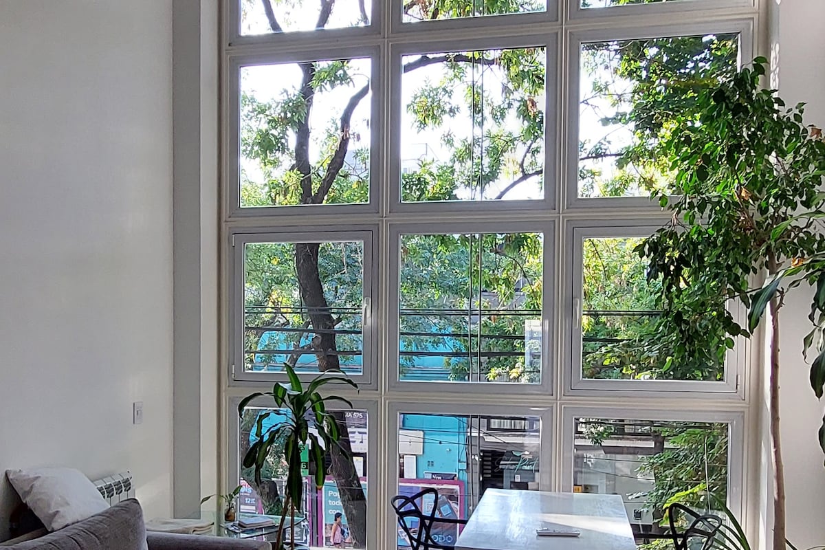 A spacious living area features large windows that allow natural light to fill the room. Greenery from outside complements the indoor plants, creating a fresh atmosphere. A simple dining table is positioned beside the windows, overlooking the vibrant street below.