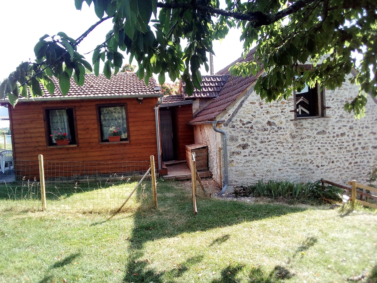 The exterior of a rustic gîte is shown, featuring a combination of wooden and stone walls. Green grass surrounds the building, with flower boxes visible beneath the windows. A wooden fence adds a boundary, while trees provide shade in the foreground.