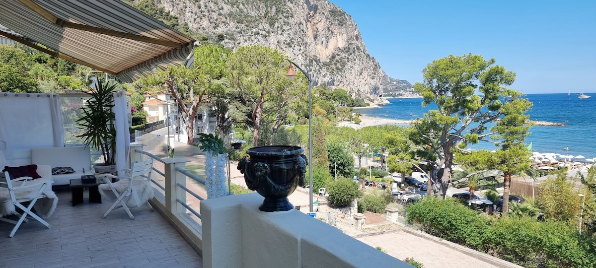 A large terrace is presented, offering sweeping views over Beaulieu beach, the port, and the surrounding landscape. Comfortable seating is arranged on the terrace, framed by green vegetation and the mountainous backdrop under a clear blue sky.