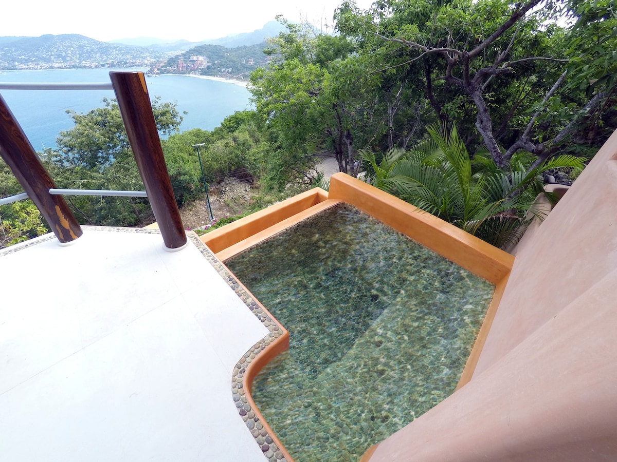 A tranquil splash pool is set on a private balcony, surrounded by natural stone accents. The serene water reflects the surrounding greenery and offers captivating views of the Bay of Zihuatanejo. The blending of outdoor and indoor spaces creates a peaceful retreat.