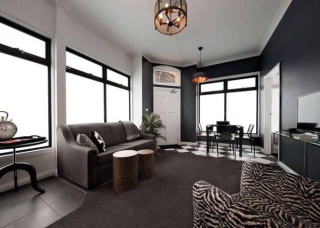 The living area features large windows that allow natural light to enhance the stylish decor. A dark sofa faces a contemporary TV unit, while a set of round tables sits beside it. A dining table is set for four, positioned near the entrance.
