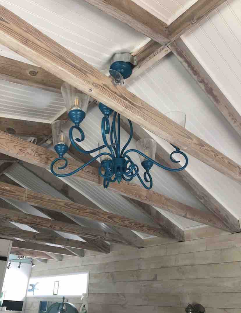 A blue chandelier with intricate swirls is suspended from a wooden-beamed ceiling. The light fixture features clear glass shades, adding a decorative touch to the space. The ceiling has a white paneling that complements the rustic wooden beams.