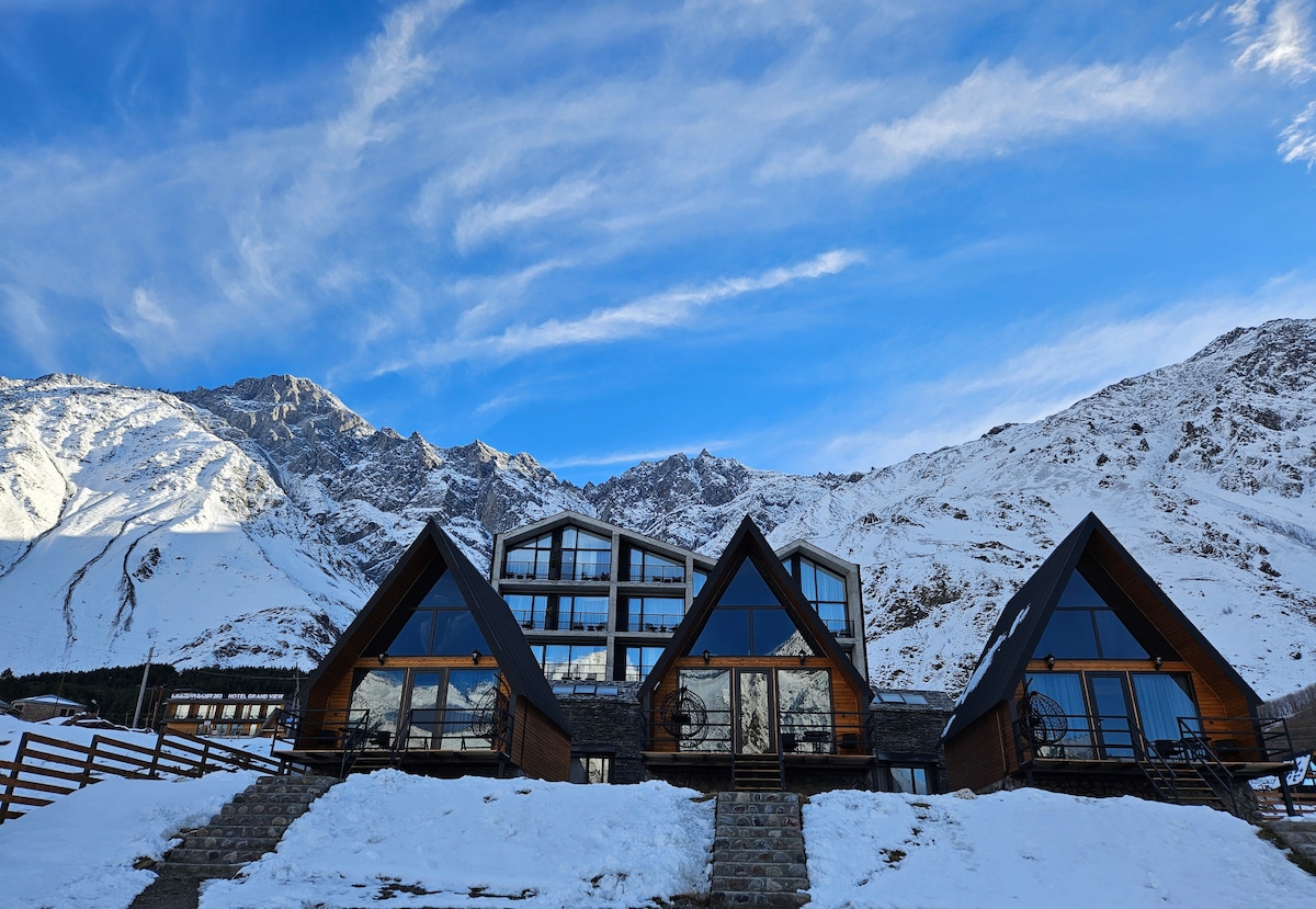 Three modern cottages with distinctive A-frame shapes are showcased against a backdrop of snow-covered mountains. Large windows reflect the clear blue sky, while a stone pathway leads up to the entrance of each unit, blending rustic architecture with the serene landscape.