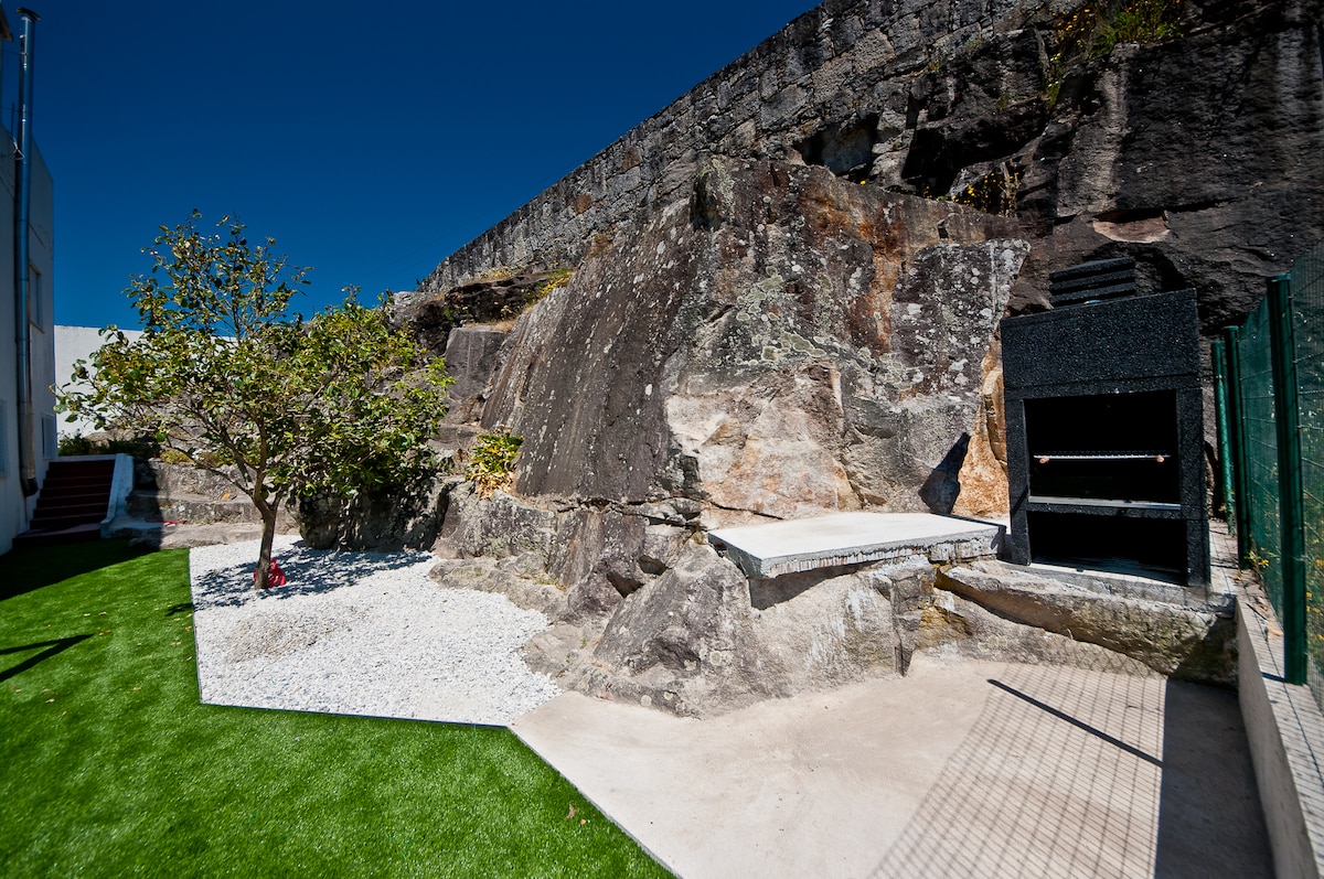 A landscaped outdoor area features a large rocky wall, with a barbecue facility positioned on a paved surface. A small tree is planted nearby, alongside a patch of gravel and a green lawn, creating a relaxed and inviting outdoor space.