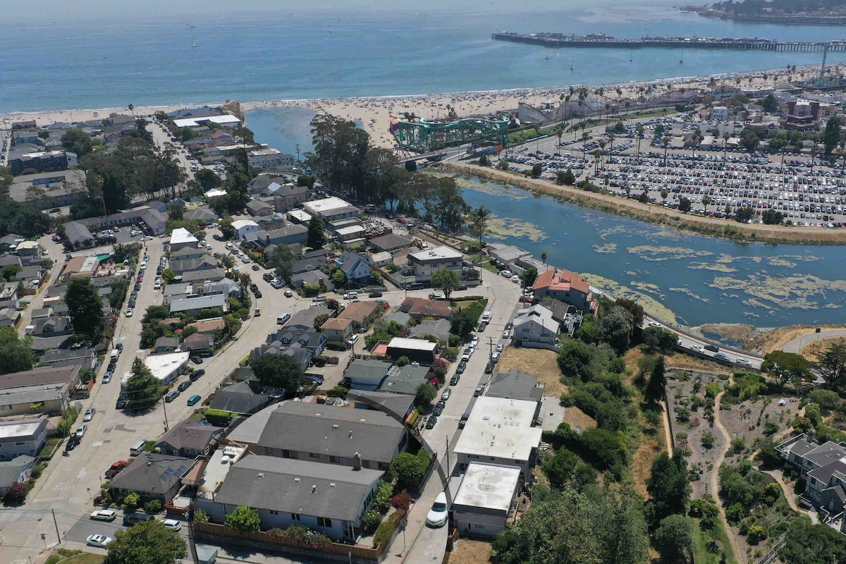An aerial view captures a coastal neighborhood featuring a mix of residential homes and winding streets. The nearby beach and boardwalk are visible, along with a marina and various parked boats along the shore. The vibrant blue ocean stretches into the distance.