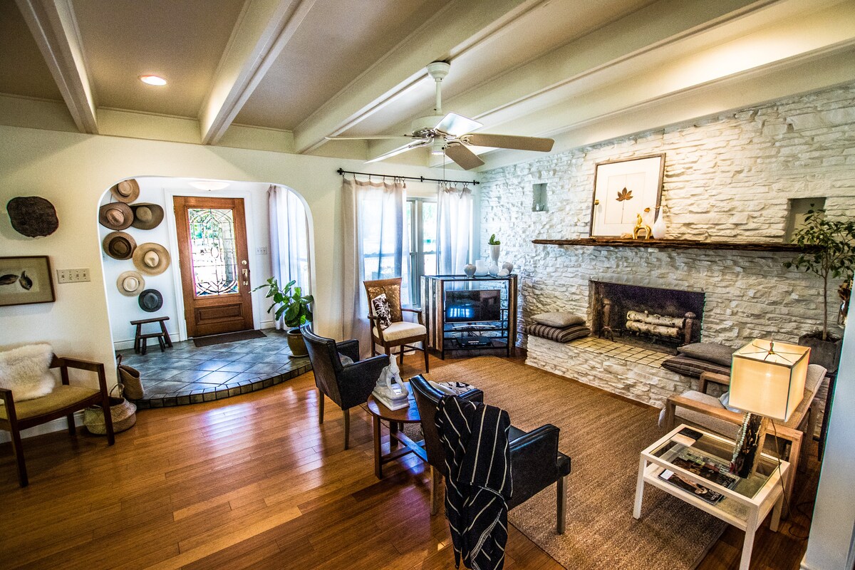 A spacious living area features natural wood floors and a stone fireplace. Comfortable seating is arranged around a coffee table, with large windows allowing ample natural light. An entrance to a tiled hallway is visible, inviting guests into the home.