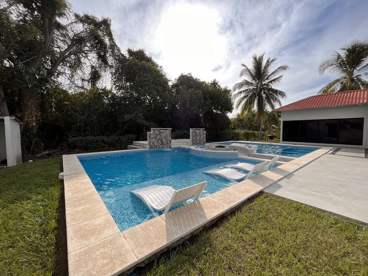 A tranquil pool area is highlighted, featuring smooth blue water and elegant lounge chairs positioned near the water's edge. Lush greenery surrounds the pool, and a modern firepit stands in the background, complemented by a nearby building with a red roof.