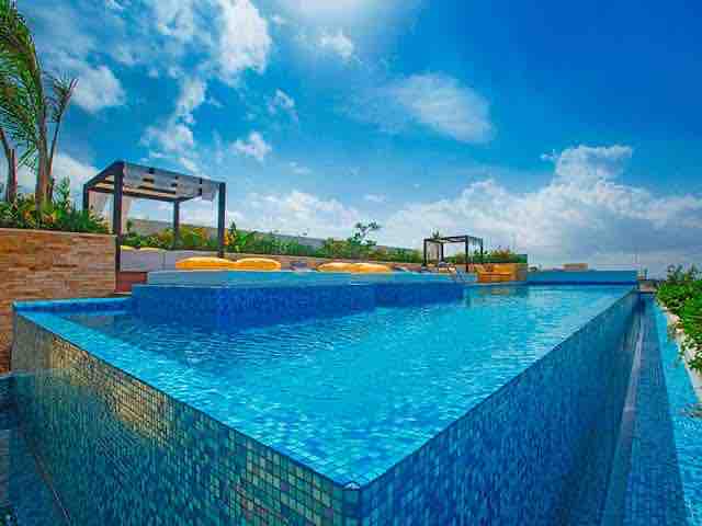 An infinity pool is showcased on a rooftop, featuring shimmering blue tiles reflecting sunlight. Comfortable lounge chairs are positioned along the pool's edge, surrounded by lush greenery. The sky is clear, with a few clouds adding depth to the vibrant scene.