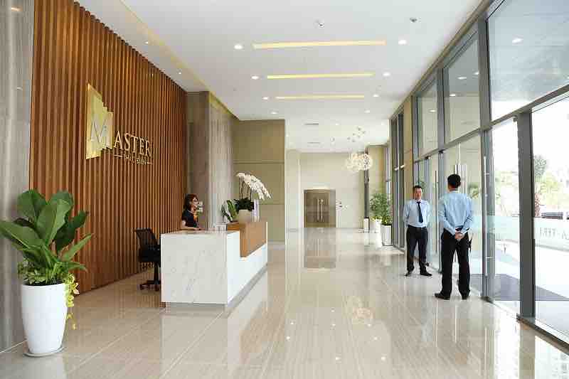 A spacious lobby area is presented with polished tile flooring and wood accent walls. A reception desk is positioned to the left, accompanied by a seating area and decorative plants. Large glass windows allow natural light, while the entrance to elevators is visible in the background.