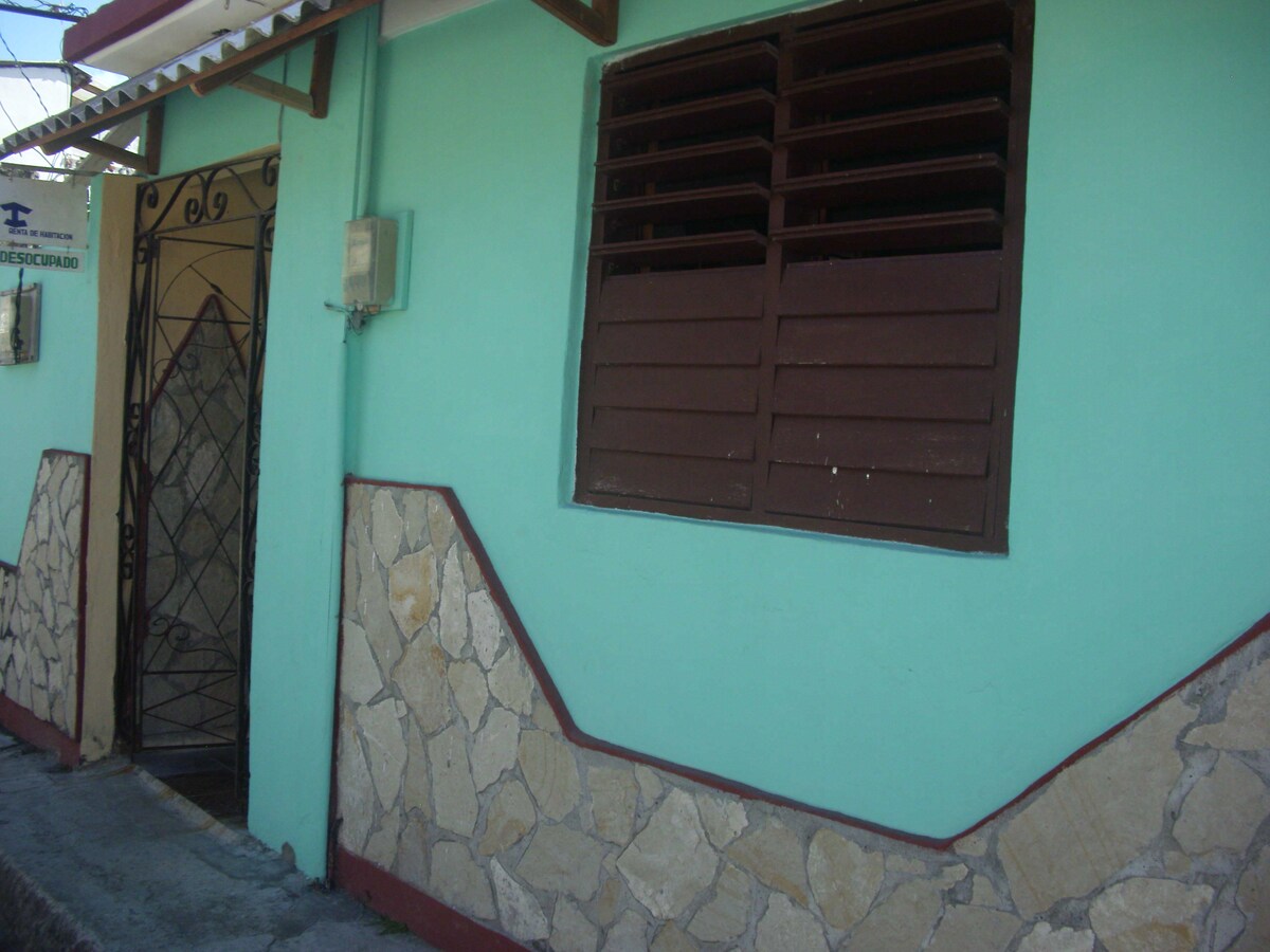 The exterior view showcases a light blue wall adorned with stone accents, featuring a wrought iron gate that leads to the entrance. A large window with wooden slats allows natural light into the space while providing ventilation.