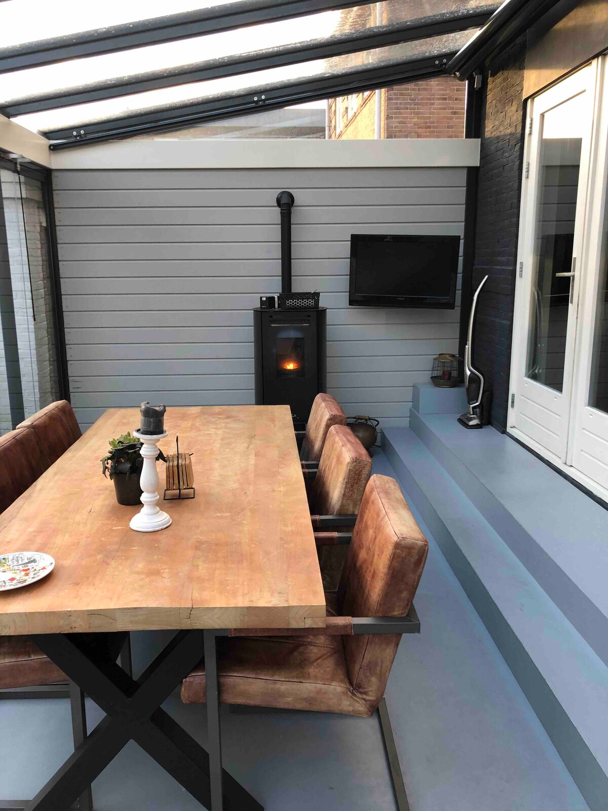 A dining area within a partially enclosed space features a wooden table surrounded by four comfortable chairs. A modern heating unit stands in the corner, while a small television is mounted on the wall. Double doors provide access to the exterior.