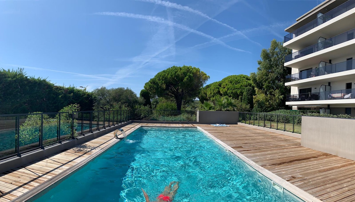 A clear swimming pool is featured surrounded by wooden decking and secured with a low metal fence. Lush greenery and trees are present in the background, complemented by a clear blue sky, enhancing the serene outdoor environment.